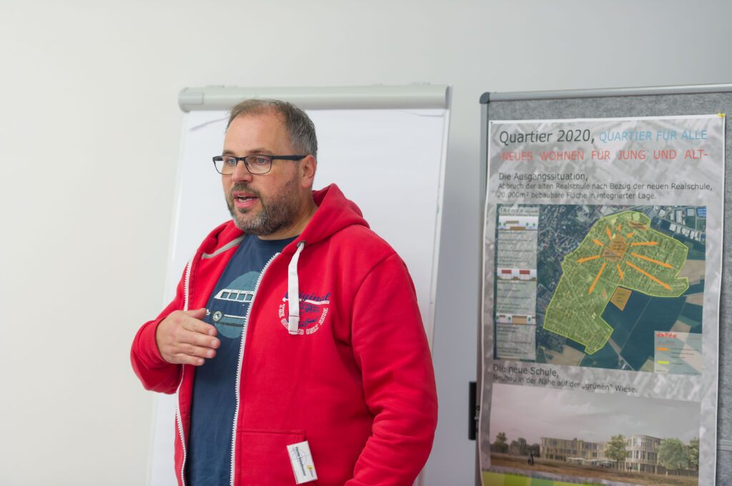 Foto im Veranstaltungsraum, ein Moderator. im roten Hoodie und Brille steht und spricht neben einem Flipchart mit Plakat bei der Veranstaltung Tatort Wohnen, Hospitalhof Stuttgart.